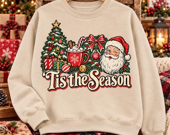 Puede incluir: Una sudadera beige de cuello redondo con un estampado gráfico de un árbol de Navidad decorado, una taza roja con un bastón de caramelo, un lazo rojo y un Santa Claus guiñando un ojo. El texto "Tis the Season" aparece en color rojo oscuro.