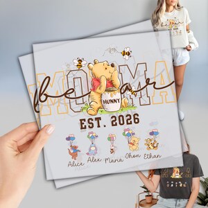 May include: A light gray print featuring Winnie the Pooh with the word "MOM" and the text "EST. 2026". Below are cartoon characters with names. The image is held by a person.