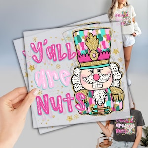 May include: A print featuring a colorful nutcracker design with the text "Y'all are Nuts" in pink. The nutcracker has a gold crown and a white mustache. The background has gold stars.