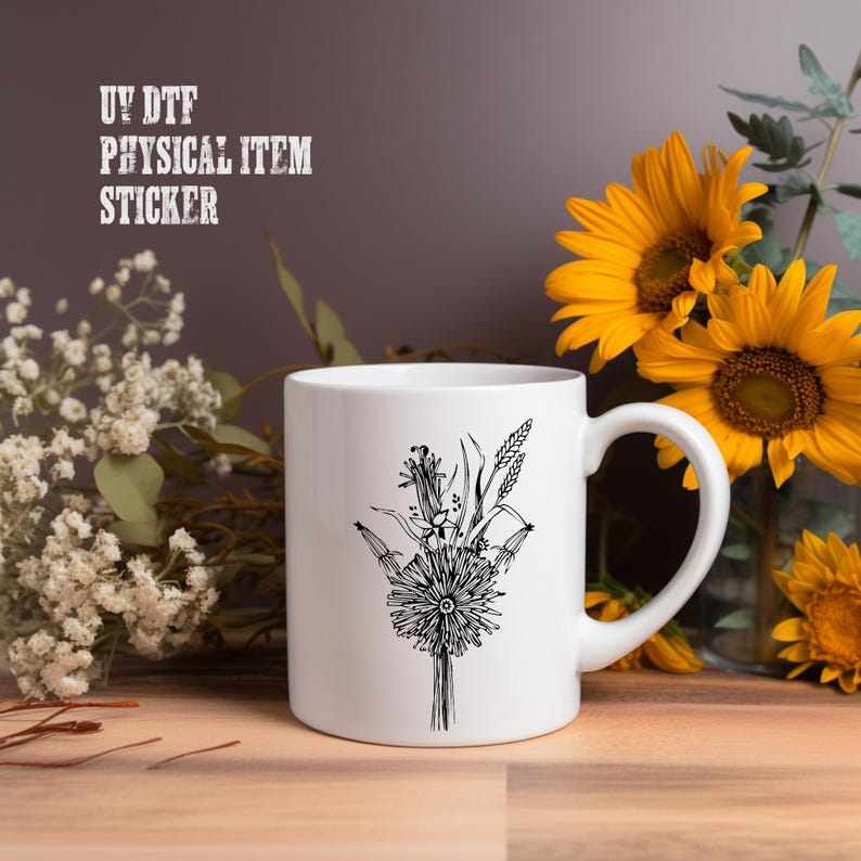 A white mug with a black floral design sits on a wooden surface, surrounded by yellow sunflowers and white flowers.