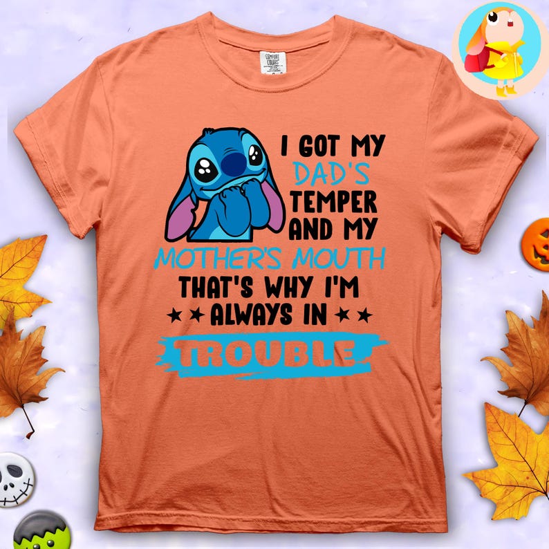Op de afbeelding: Oranje T-shirt met een blauw stripfiguur en de tekst "I GOT MY DAD'S TEMPER AND MY MOTHER'S MOUTH THAT'S WHY I'M ALWAYS IN TROUBLE". Het shirt heeft korte mouwen en een ronde hals.