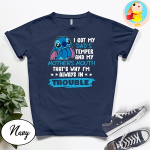 Op de afbeelding: Marineblauw T-shirt met een cartoonfiguur en de tekst "I GOT MY DAD'S TEMPER AND MY MOTHER'S MOUTH THAT'S WHY I'M ALWAYS IN TROUBLE." Het ontwerp bevat blauwe en witte accenten. Een paar sneakers is zichtbaar.