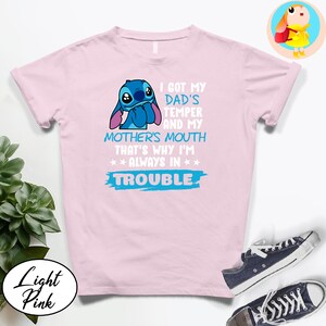 Op de afbeelding: Lichtroze T-shirt met een blauw stripfiguur en de tekst "I GOT MY DAD'S TEMPER AND MY MOTHER'S MOUTH THAT'S WHY I'M ALWAYS IN TROUBLE". Het shirt wordt gecombineerd met blauwe sneakers.