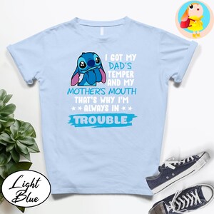 Op de afbeelding: Lichtblauw T-shirt met een Stitch-afbeelding en de tekst "I GOT MY DAD'S TEMPER AND MY MOTHER'S MOUTH THAT'S WHY I'M ALWAYS IN TROUBLE". Het shirt heeft ook de woorden "Light Blue" in een cirkel.