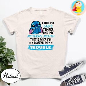 Op de afbeelding: Een wit t-shirt met een blauw cartoonfiguur en de tekst "I GOT MY DAD'S TEMPER AND MY MOTHER'S MOUTH THAT'S WHY I'M ALWAYS IN TROUBLE." Het shirt wordt gecombineerd met marineblauwe sneakers.