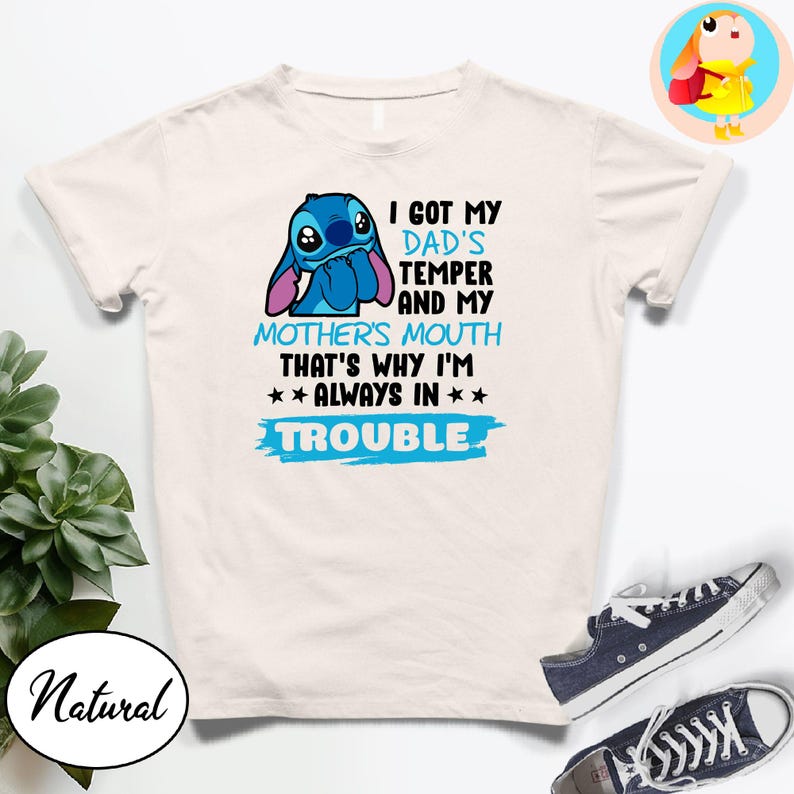 Op de afbeelding: Een wit t-shirt met een blauw cartoonfiguur en de tekst "I GOT MY DAD'S TEMPER AND MY MOTHER'S MOUTH THAT'S WHY I'M ALWAYS IN TROUBLE." Het shirt wordt gecombineerd met marineblauwe sneakers.