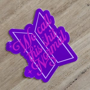 May include: A purple sticker in a star shape with the text "We call this shit Normal" in a pink, cursive font. The sticker has a white border and is on a wooden surface.