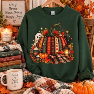 May include: A dark green crewneck sweatshirt featuring a graphic of a large pumpkin made of stacked books, a small white ghost, a black cat, a lantern, and autumn leaves. The design includes a plaid blanket and two mugs with pumpkin motifs.