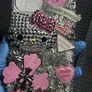 Puede incluir: Funda de teléfono transparente adornada con pedrería, con un diseño de Hello Kitty, un lazo rosa y varios adornos plateados y rosas. La funda incluye el nombre "EVA" y un charm en forma de corazón.