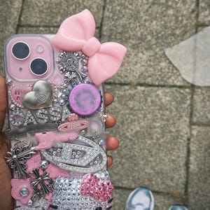 May include: Pink and silver embellished phone case with a large pink bow, silver charms, and a Hello Kitty design. The case features rhinestones, crosses, and the word "CASSA". The phone case is a decorative accessory.