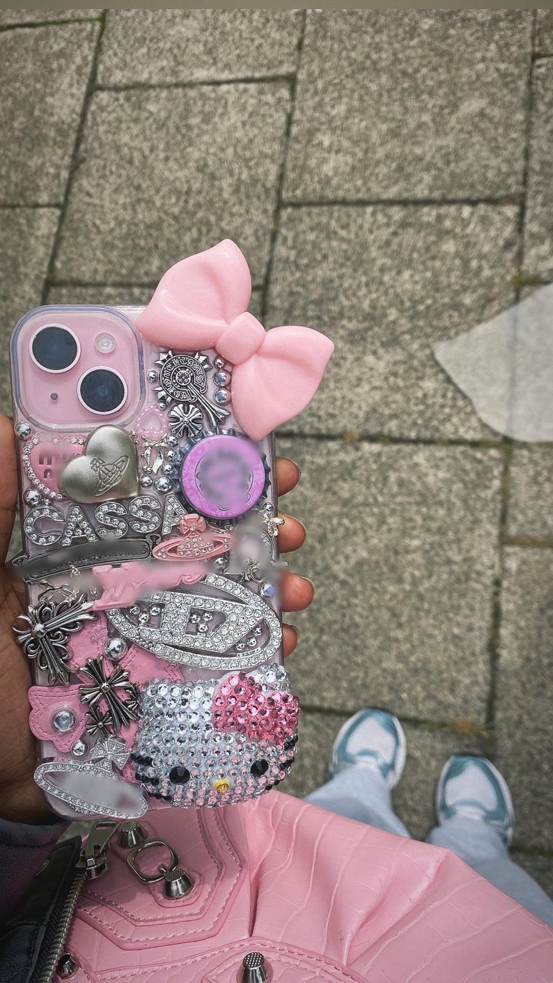 May include: Pink and silver embellished phone case with a large pink bow, silver charms, and a Hello Kitty design. The case features rhinestones, crosses, and the word "CASSA". The phone case is a decorative accessory.
