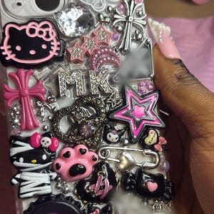Può includere: Custodia per telefono trasparente decorata con vari ciondoli, tra cui un Hello Kitty rosa, stelle, croci e un'impronta di zampa. La custodia ha una varietà di colori, tra cui rosa, nero, argento e bianco. La custodia è impreziosita da strass e altri elementi decorativi.
