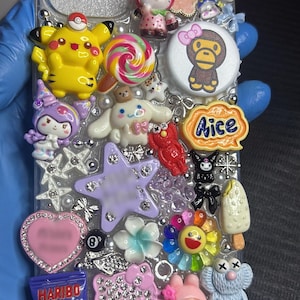 May include: A clear phone case decorated with various colourful 3D charms including Pikachu, Hello Kitty, a lollipop, a Haribo Stamix candy bag, a flower, a star, a heart, and other cartoon figures, all arranged on a glittery, textured surface.