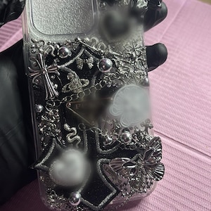 May include: Clear phone case with black and silver embellishments. Features a black bow, crosses, a butterfly, and a snake. The case is decorated with silver chains and beads, creating a gothic aesthetic.