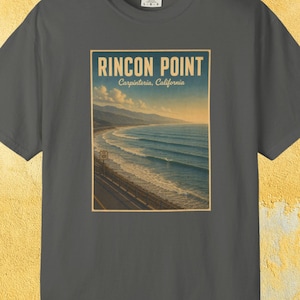May include: Dark gray t-shirt featuring a rectangular graphic of Rincon Point in Carpinteria, California. The graphic depicts a coastal landscape with ocean waves, a road, and the text "Rincon Point". The shirt has a crew neck and short sleeves.