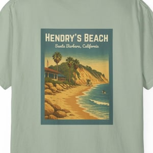 May include: A light green t-shirt featuring a vintage-style illustration of Hendry's Beach in Santa Barbara, California. The design includes a beach scene with palm trees, a lifeguard tower, and the text "HENDRY'S BEACH".