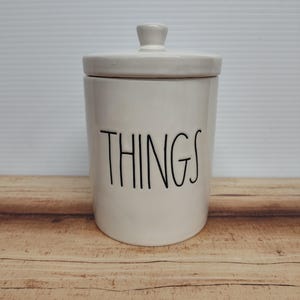 May include: A cream-colored ceramic jar with a lid and a small knob. The word "THINGS" is written in black, handwritten-style lettering on the front of the jar. The jar is sitting on a wooden surface.