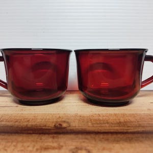May include: Two ruby red glass mugs with handles. The mugs have a rounded shape and a dark rim. They are sitting on a wooden surface against a white background. The mugs are empty.