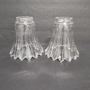 May include: Two clear glass lamp shades with a fluted design. The shades have a bell shape with a scalloped edge. The shades are made of clear glass and are sitting on a dark surface.