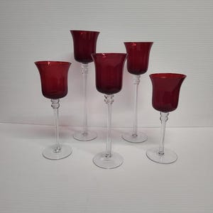 May include: Five ruby red glass candle holders. Each holder has a flared top, a long, clear stem, and a round base. The candle holders are arranged in a group, showcasing their elegant design.