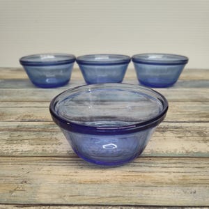 May include: Four blue glass bowls. The bowls are a light blue color and have a rounded shape with a slightly flared rim. The bowls are arranged on a wooden surface.