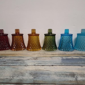 May include: Six colorful glass candle holders in shades of red, amber, yellow, green, and blue. Each holder has a diamond pattern and a small, square top. The candle holders are arranged in a row on a wooden surface.