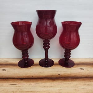 May include: Three ruby red glass goblets with a bulbous bowl and a decorative stem. The goblets are arranged on a wooden surface, with a white background. The central goblet is taller than the two flanking it.