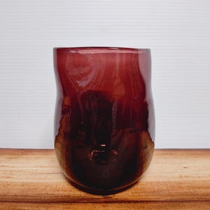 May include: A deep burgundy-colored glass vase with a rounded, organic shape. The vase has a smooth, glossy finish and sits on a wooden surface. The glass appears to have small bubbles within the material, adding texture.