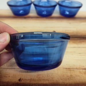 May include: Three cobalt blue glass bowls. The bowls have a rounded shape with a slightly flared rim. The glass has a smooth, glossy finish. The bowls are arranged on a wooden surface.