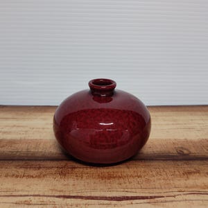 May include: A round, burgundy-colored ceramic vase with a speckled texture. The vase has a small, cylindrical opening at the top and sits on a wooden surface. The vase is a decorative item.