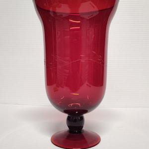 May include: A tall, ruby red glass vase with a flared top and a round base. The vase has a bulbous midsection and a dark red stem. Delicate etched lines adorn the side of the vase. The vase is likely used for decorative purposes.