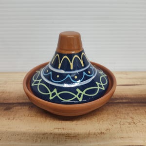 May include: A ceramic tagine pot with a brown base and lid. The lid is dark blue with yellow and white decorative patterns. The base is a lighter brown color. The tagine is on a wooden surface.