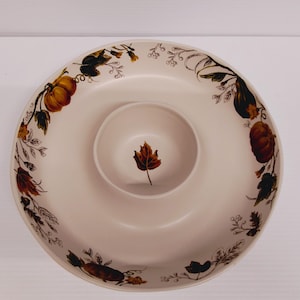 May include: Cream-colored serving platter with a central dip bowl. The rim is decorated with pumpkins, leaves, and vines in shades of orange, brown, and green. A single leaf is painted in the center of the dip bowl. Ideal for fall gatherings.