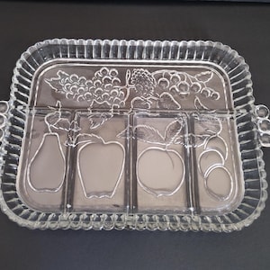 May include: Clear glass serving tray with a rectangular shape and four divided sections. The tray features embossed fruit designs, including grapes, pears, apples, and peaches. The edges have a ribbed design, and the tray has rounded handles.