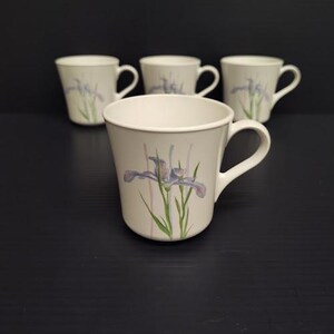 May include: A set of four off-white ceramic mugs, each featuring a delicate watercolor-style illustration of a purple iris flower with green stems and leaves. The mugs have a flared rim, a curved handle, and a smooth, glossy finish throughout.