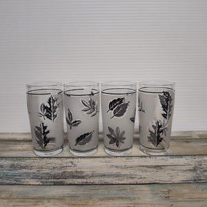 May include: Four clear drinking glasses with a frosted leaf design in black and gray. The glasses are cylindrical and sit on a weathered wooden surface. The leaf patterns vary on each glass, adding a unique touch to the set.