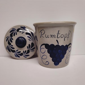 May include: A light gray ceramic crock with a matching lid, featuring a dark blue hand-painted grape cluster and vine motif. The word Rumtopf is written in dark blue script on the front of the vessel, which has a cylindrical shape and a knobbed lid.