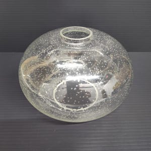 May include: A clear, spherical glass vase featuring a textured surface with numerous small, white, bubble-like inclusions throughout the body. The vase has a circular opening at the top with a smooth, reinforced rim, creating a rounded silhouette.