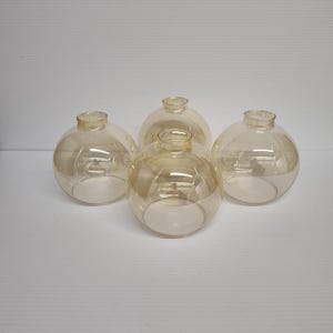 May include: Four clear, round glass globes with a light amber tint. Each globe has a small, open neck and a spherical body. The glass appears smooth and reflective, suitable for lighting fixtures or decorative purposes.