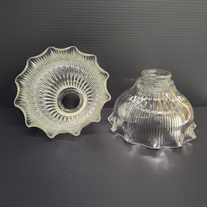 May include: Two clear glass lamp shades. One is a scalloped, open-faced design, and the other is a bell-shaped shade with a ribbed texture. Both are made of clear glass.