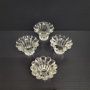 May include: Four clear glass candle holders with a scalloped, seashell-like design. The holders are arranged on a dark surface, showcasing their intricate details and reflective qualities. These decorative pieces add a touch of elegance.