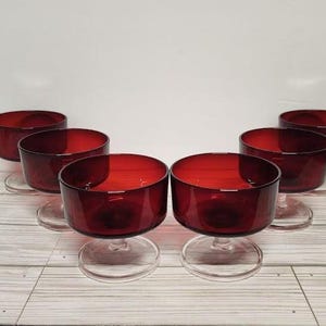 May include: Six vintage ruby red glass dessert bowls with clear stems and bases. The bowls are arranged on a light wood-grain surface. The bowls have a rounded shape and are made of a transparent glass.