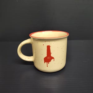 May include: Cream-colored ceramic mug with a red rim and handle. The mug features a red graphic of a stylized object with drips. The mug has a speckled appearance and is set against a dark background.