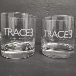 May include: Two clear, cylindrical drinking glasses with the word "TRACE3" etched in white. The glasses are made of transparent glass and sit on a dark surface. The glasses are likely used for drinking beverages.