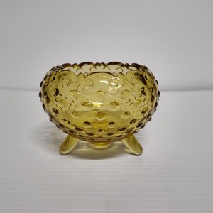 May include: Amber-colored glass bowl with a scalloped rim and a textured, bubble-like pattern. The bowl is supported by three small, curved feet. The glass has a warm, golden hue, and the bowl is likely vintage or antique.