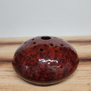 May include: A round, red ceramic vase with a speckled texture and multiple small holes on top. The vase has a glossy finish and a single larger opening in the center. It sits on a wooden surface.
