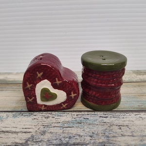 May include: A pair of ceramic salt and pepper shakers. One is heart-shaped, with a red exterior and white and green heart details. The other is spool-shaped, with a red, textured body and a green top and base. The shakers are on a wooden surface.