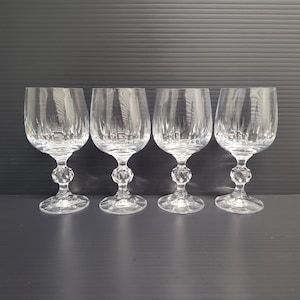 May include: Four clear crystal wine glasses with a decorative stem and a patterned bowl. The glasses are arranged in a row against a dark background. The stems have a diamond-like design.