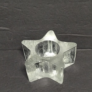 May include: A clear glass star-shaped candle holder. The candle holder has a textured surface and a circular indentation in the center to hold a candle. The star has five points and is sitting on a dark surface.