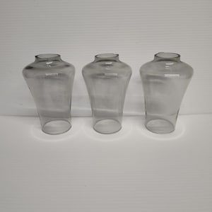 May include: Three clear glass lamp shades with a flared, vase-like shape. Each shade has a rounded top and a wide, open base. The shades are arranged in a row against a plain white background. The glass appears smooth and transparent.